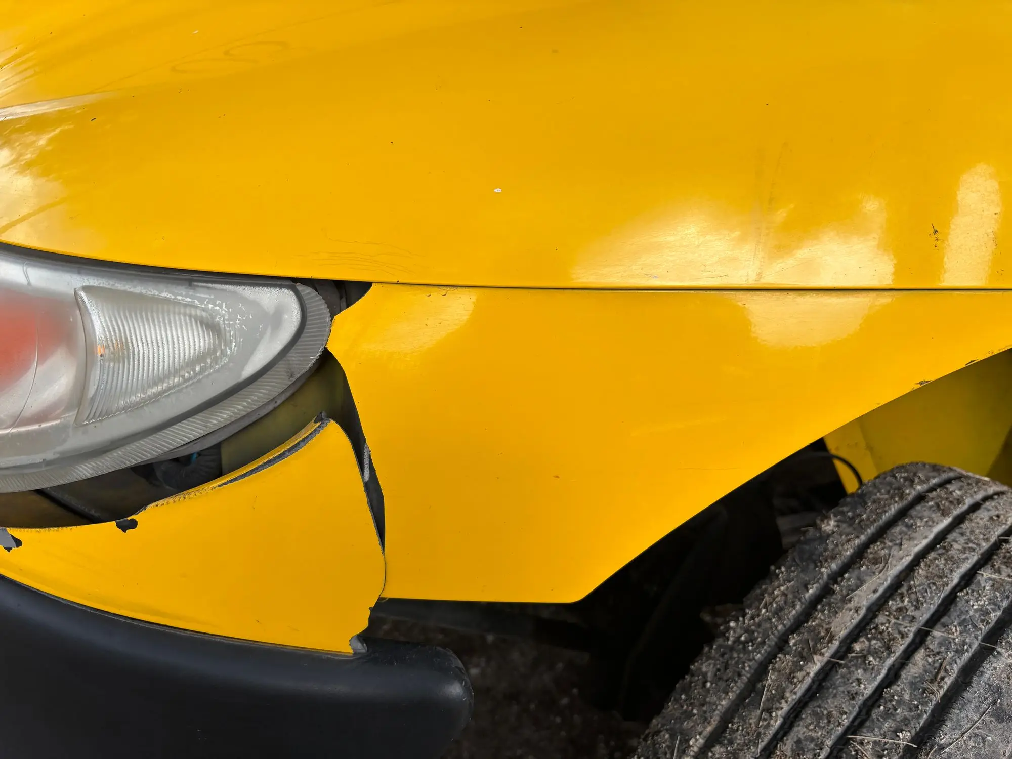 Close-up of a crumpled front guard and cracked headlight surround on a yellow work vehicle after a collision