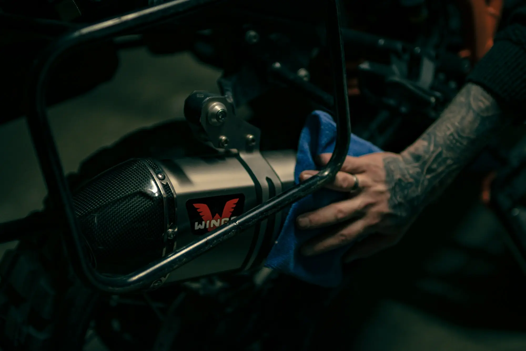Close-up of a hand wiping down a vehicle's exhaust with a blue cloth in a dim workshop, the kind of final check done before a vehicle is handed over
